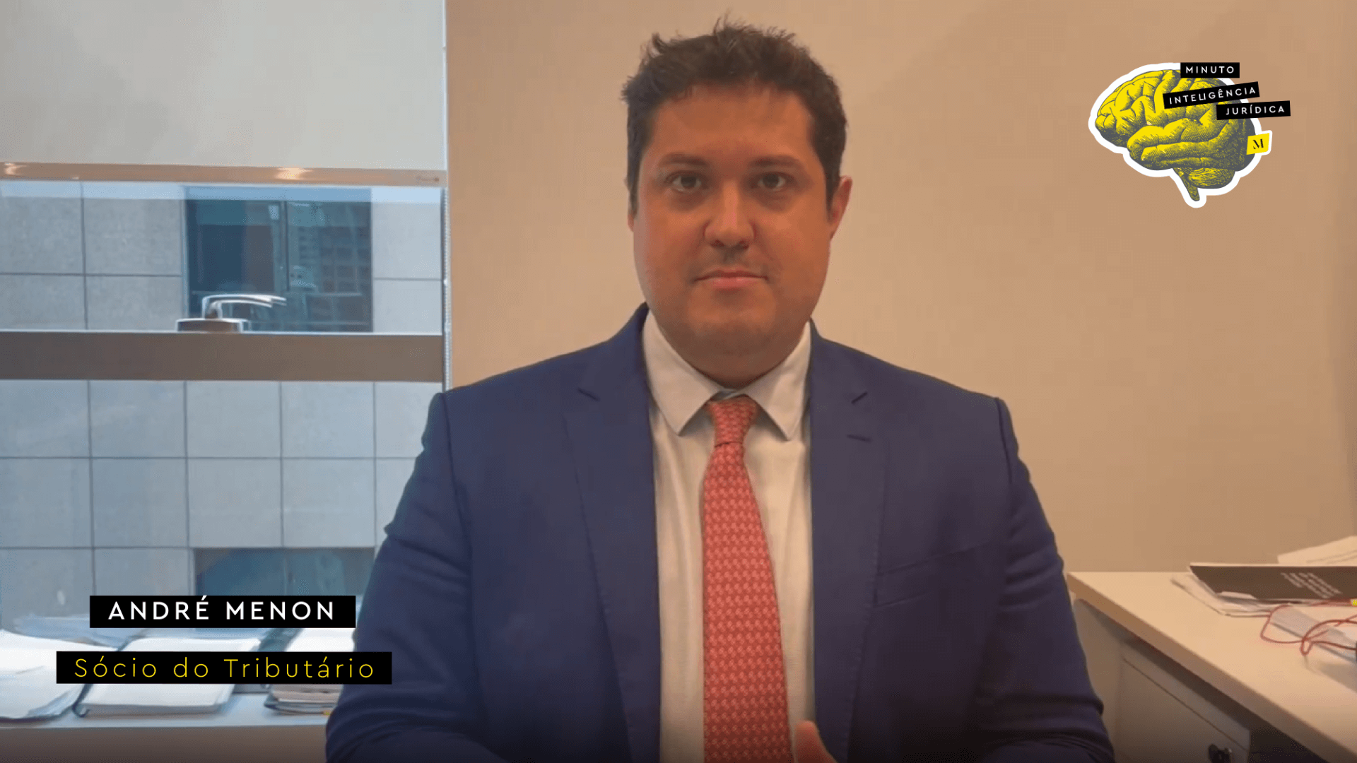Homem com cabelo preto, usando camisa branca, gravata salmão com bolinhas brancas e blazer azul marinho. No canto inferior esquerdo, faixa preta com frase nas cores branco e amarelo escrito: André Menon, sócio do Tributário. No canto superior direito, imagem ilustrativa de cérebro amarelo com faixa de texto escrito: Minuto Inteligência Jurídica