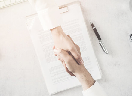 Duas pessoas brancas dando as mãos como forma de "acordo". Ao fundo da imagem, contrato assinado pelas partes em cima de uma mesa