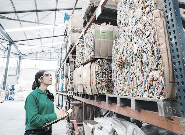 A woman wearing a green T-shirt, goggles and her hair in a ponytail, holding a clipboard and looking at a stock of materials in a factory